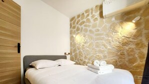 A neatly made bed with white linens and two rolled towels, a stone wall with a warm light, and a wooden door.