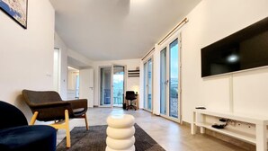 A modern living room with a sofa, a chair, a white ottoman, a TV, and a balcony with sliding doors.