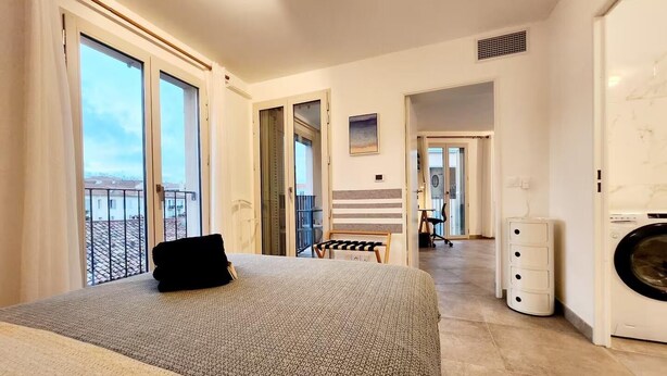 A bedroom with a bed, a washing machine, and a balcony with a view of buildings.