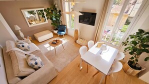A modern living room with a sofa, coffee table, and dining table. There is a large window providing natural light and a view of the outside. - Le Delmas Riviera - Duplex dans Vieil Antibes (Antibes)