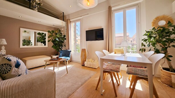 A spacious living room with a sofa, a dining table with chairs, a large plant, and a TV mounted on the wall. - Le Delmas Riviera - Duplex dans Vieil Antibes (Antibes)