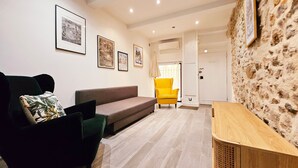 A modern living room with a sofa, armchair, and a wooden side table. There are framed pictures on the wall and a stone wall on one side.