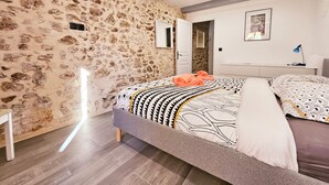 A bedroom with a stone wall, a bed with patterned bedding, a bedside table with a lamp, and a framed picture on the wall.