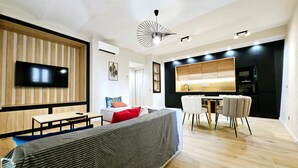 A modern living room with a large TV, a sofa, a dining table, and a kitchen area.