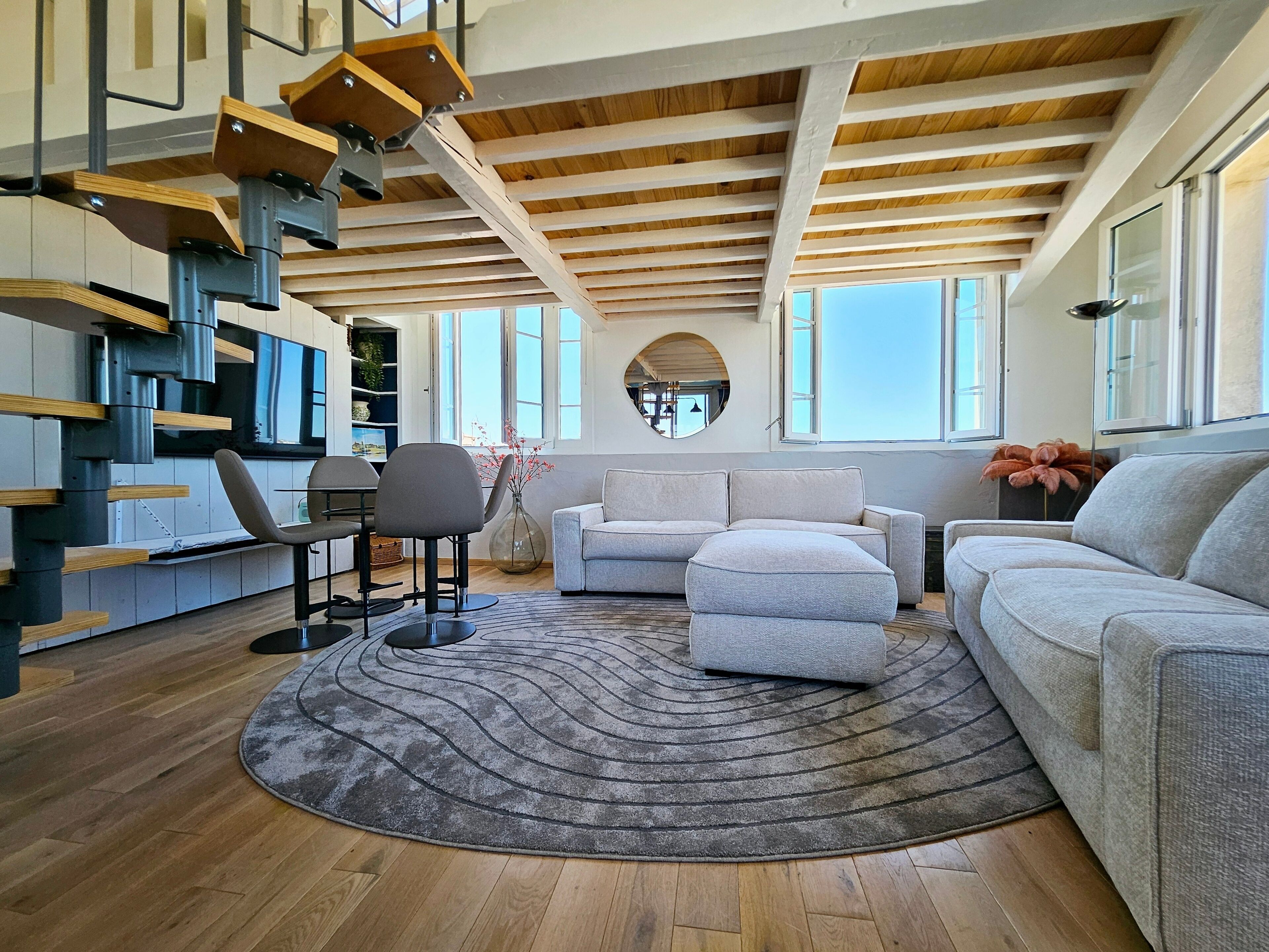 A modern living room with a wooden ceiling, a grey sofa, a round rug, and a dining area with a chair and table. - Le Phare Azur - T3 avec Vue Mer (Antibes)