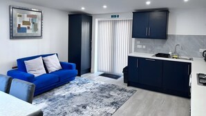 Un salon moderne avec un canapé bleu, un coin cuisine doté de meubles sombres et une grande fenêtre avec des stores.