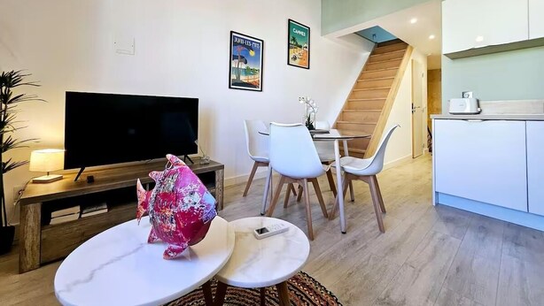 A modern living room with a flat-screen TV, a dining area with white chairs, a staircase, and a kitchen area with white cabinets.