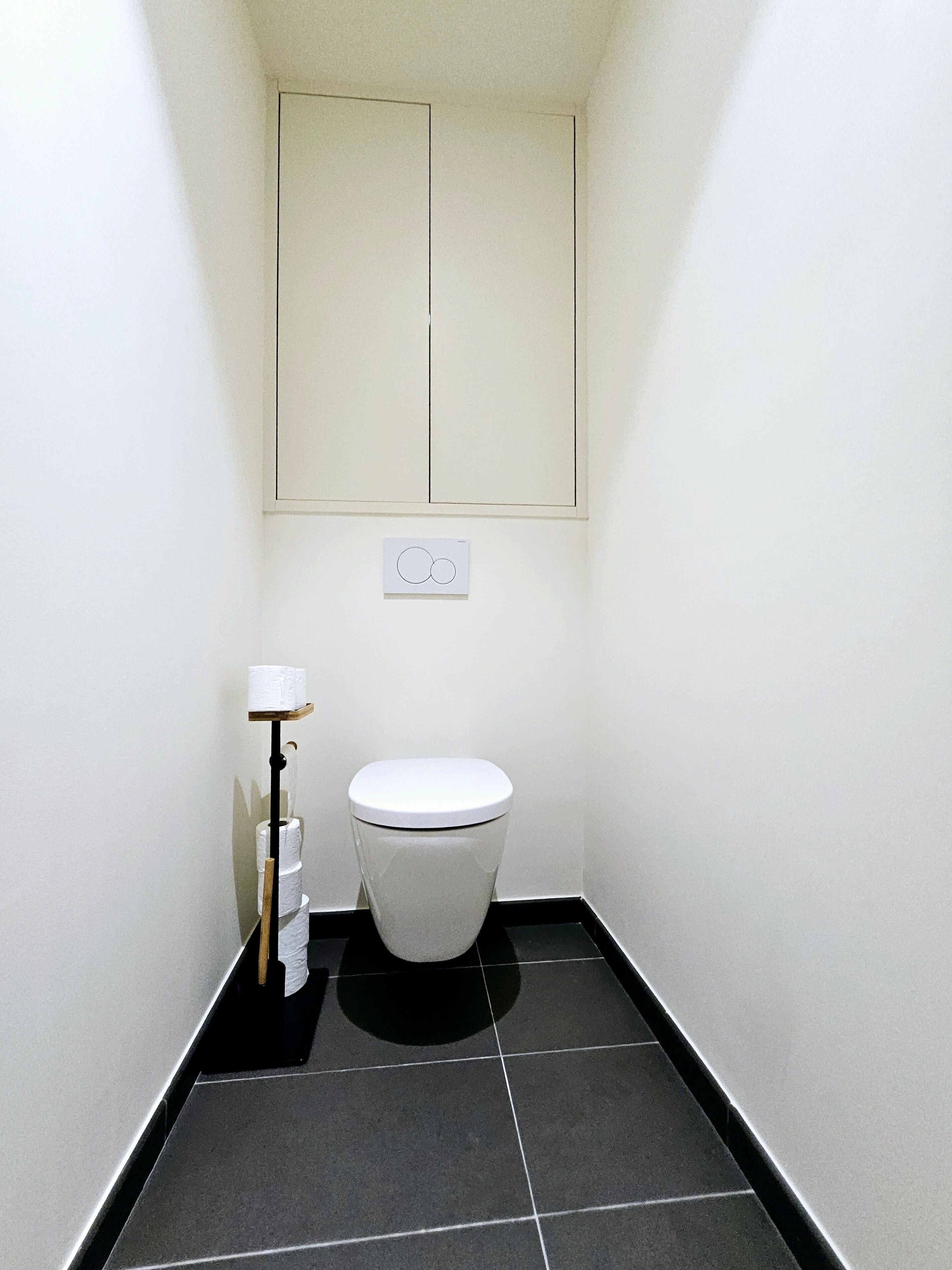 A small, narrow bathroom with a white toilet, a toilet paper holder, and a wall-mounted light switch.