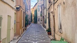 Une rue étroite, bordée de bâtiments des deux côtés, avec un trottoir à motifs et du mobilier de jardin vert.