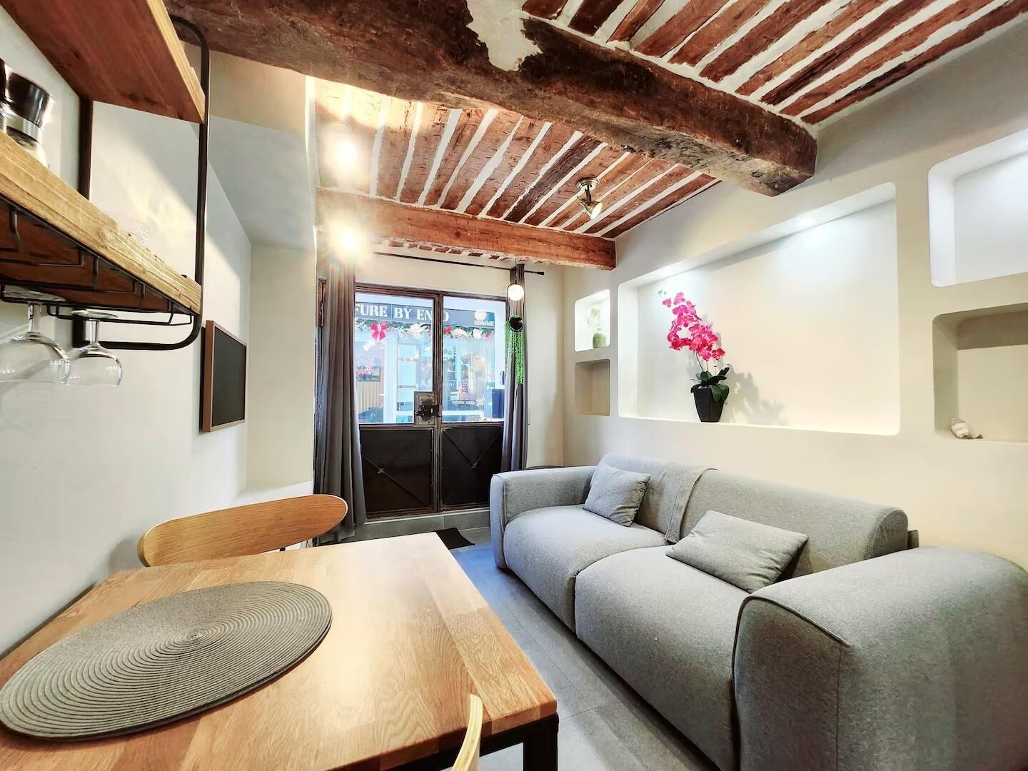 A cozy room with a wooden ceiling, a grey sofa, a wooden table, and a window with a view of the outside.