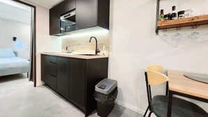 Une cuisine compacte avec des meubles de rangement sombres, un four à micro-ondes encastré et un petit évier. On y trouve une poubelle et un coin repas avec une table et des chaises.
