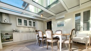 A modern kitchen with white cabinets, a dining area with wooden chairs, and a large window providing natural light.