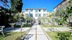 Un chemin mène à un bâtiment blanc aux volets verts, entouré de verdure et d’un jardin.