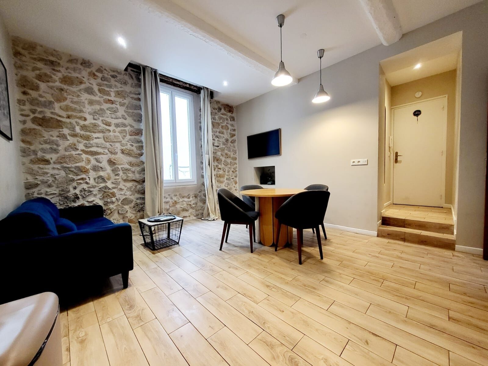 A living room with a stone wall, a wooden floor, a sofa, a coffee table, a dining table with chairs, and a TV mounted on the wall.