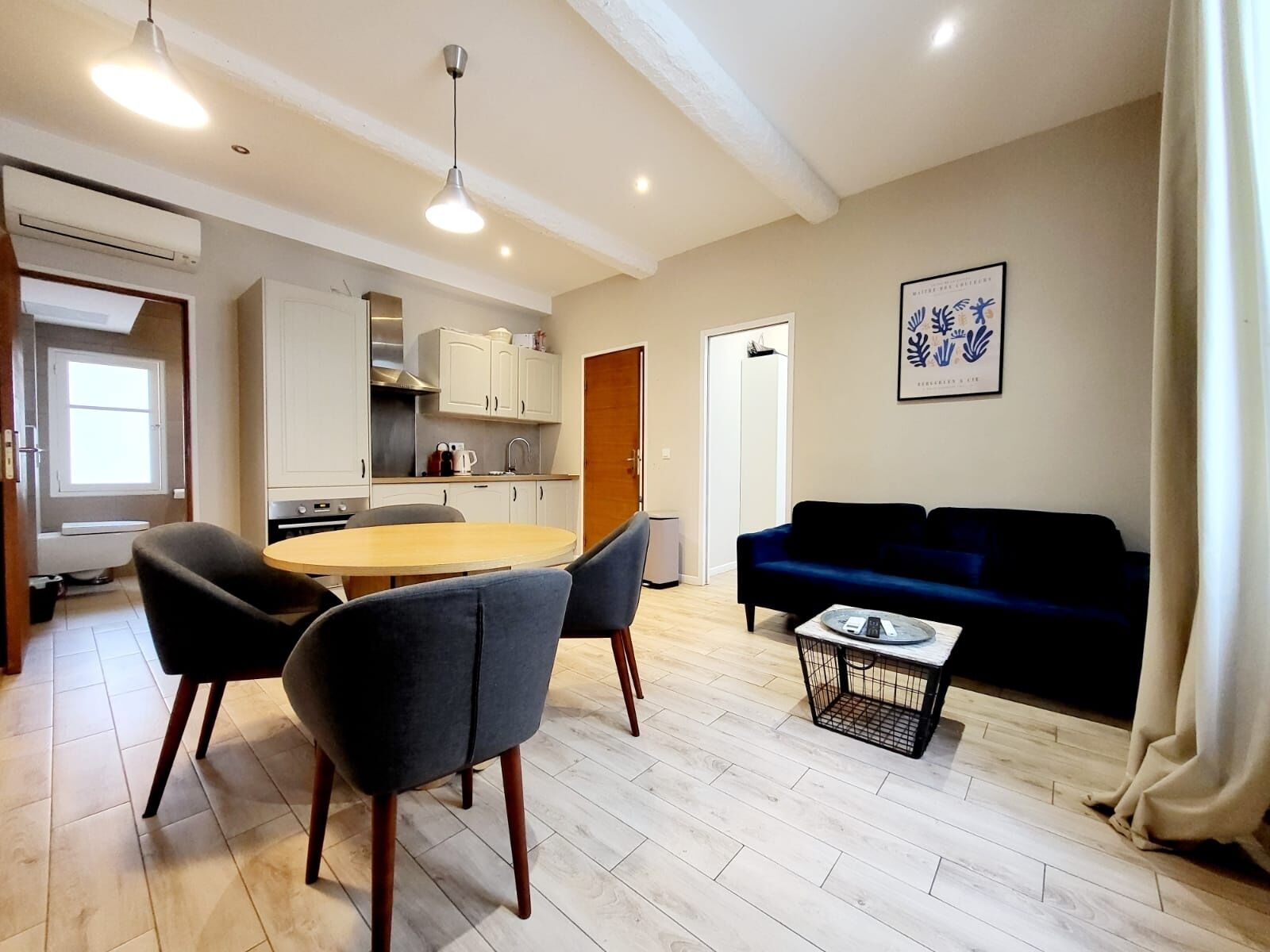 A modern living room with a dark blue sofa, a round wooden dining table, and two grey chairs. There is a kitchen area with white cabinets and a window.