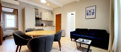 A modern living room with a dark blue sofa, a round wooden dining table, and two grey chairs. There is a kitchen area with white cabinets and a window.
