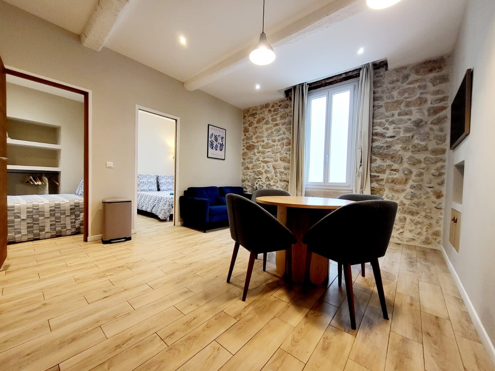 A cozy room with a wooden floor, a dining table with chairs, a sofa, a bed with patterned bedding, a stone wall, and a window with curtains.