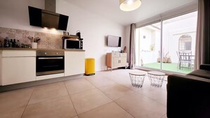 A modern kitchen with a microwave, oven, and a yellow trash bin. There is a dining area with a table and chairs, and a large sliding glass door leading to an outdoor space. - Le Thuret Fleuri - T3 avec Terrasse (Antibes)