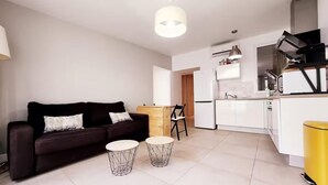 A modern living room with a dark sofa, two wireframe tables, and a kitchen area with white cabinets and a yellow trash bin. - Le Thuret Fleuri - T3 avec Terrasse (Antibes)
