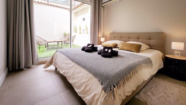 A bedroom with a bed, two bedside lamps, a window with curtains, and an air conditioning unit. - Le Thuret Fleuri - T3 avec Terrasse (Antibes)