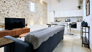 A modern living room with a stone wall, a flat-screen TV, a sofa, and a kitchen area with white cabinets and a countertop.