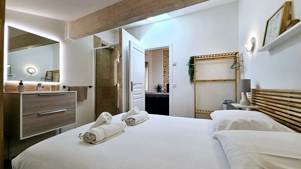 A modern hotel room with a large bed, a wooden headboard, a bedside table with a lamp, a mirror, and a bathroom with a shower and a sink.
