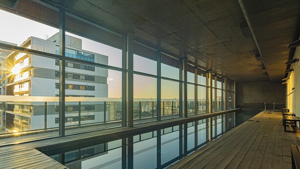 Un área de piscina interior moderna con amplios ventanales, suelo de madera y vistas a un paisaje urbano.