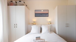 A neatly made bed with a folded towel and a card, white wardrobe, and a painting of a sunset over the ocean.