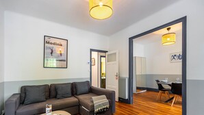 A modern living room with a grey sectional sofa, a small round coffee table, and a white ottoman. There is a framed poster on the wall and a dining area with a table and chairs visible through an open doorway.
