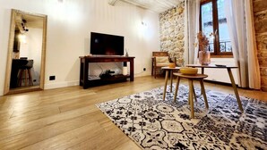 A living room with a TV on a wooden stand, a stone wall, a window with curtains, and a patterned rug.