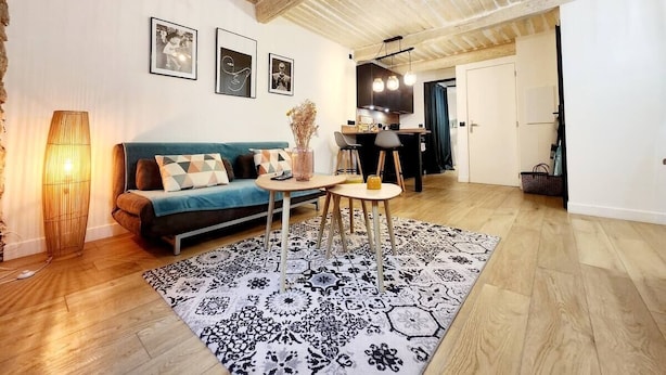 A living room with a sofa, coffee table, and patterned rug. There is a dining area with a table and chairs. The room has wooden flooring and a ceiling with exposed beams.
