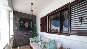 Ein Balkon mit Steinmauer, Dartscheibe, einer Couch mit tropischen Kissen und einem Fenster mit Jalousien.