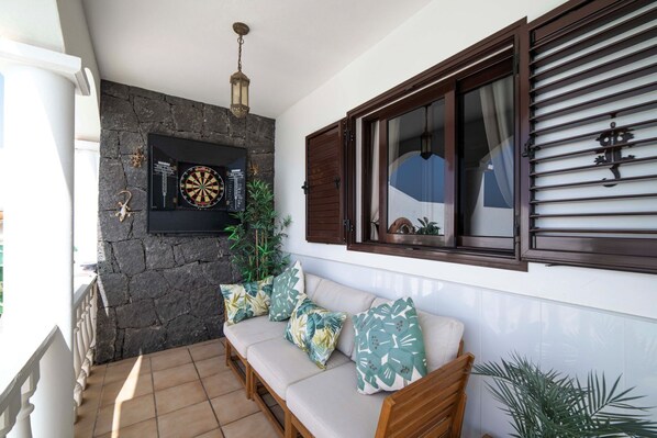 Un balcón con muro de piedra, dardo, sofá con cojines tropicales y ventana con persianas.