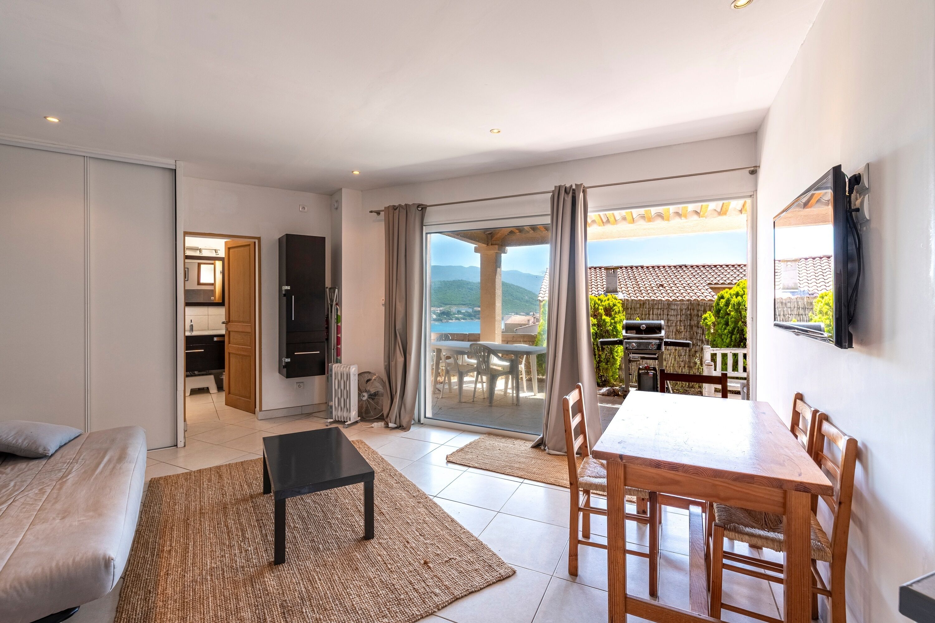 A modern living room with a sofa, a dining table with chairs, a black coffee table, and a view of a balcony with outdoor furniture.