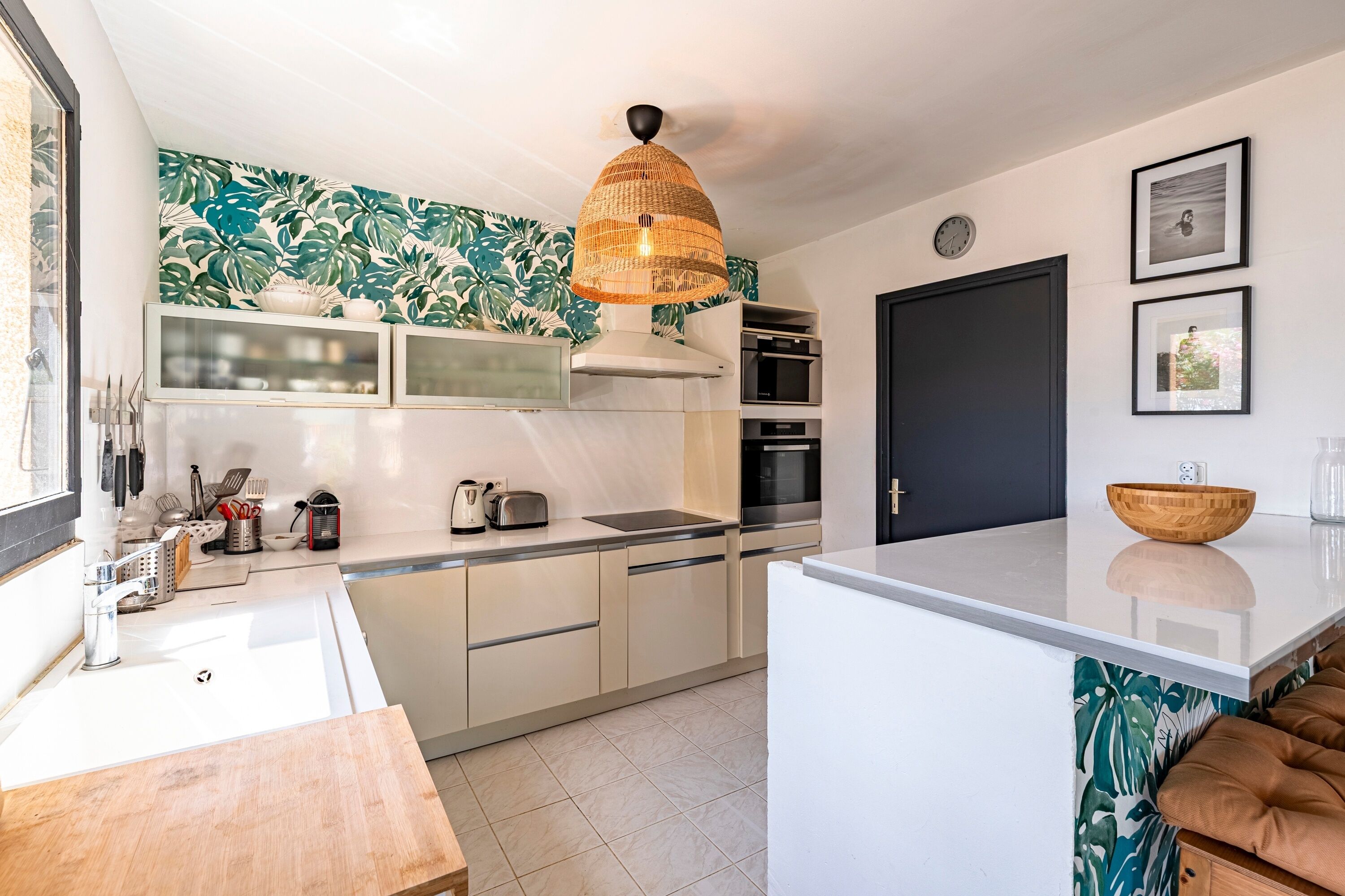 Een moderne keuken met witte kasten, een groot eiland, een hangende lamp en een spatwand met een tropisch bladmotief.