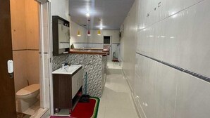 A narrow hallway with a bathroom on the left, a kitchenette area in the center, and a tiled wall on the right.