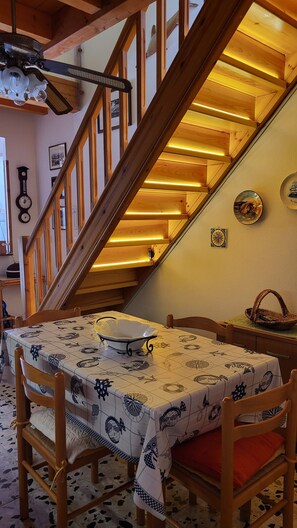 Ein Essbereich mit einem Tisch für vier Personen, Holzstühle, einer gemusterten Tischdecke und einer Treppe mit Holzwangen, die in ein Obergeschoss führt.