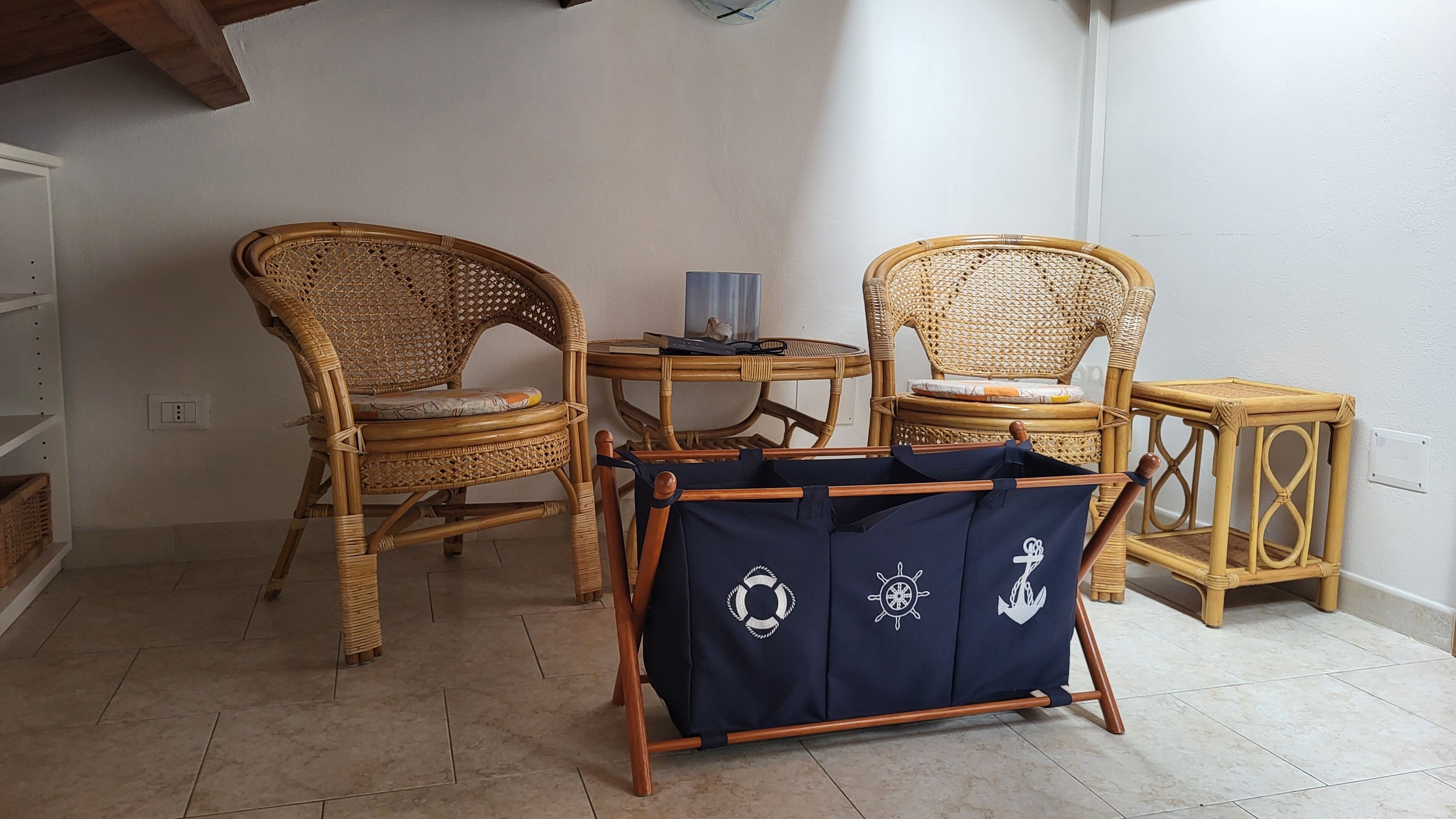 Ein Zimmer mit zwei Korbstühlen, einem kleinen runden Tisch mit einem Buch und einem dekorativen Gegenstand, sowie einer marineblauen Tasche mit maritimen Symbolen auf einem Ständer.