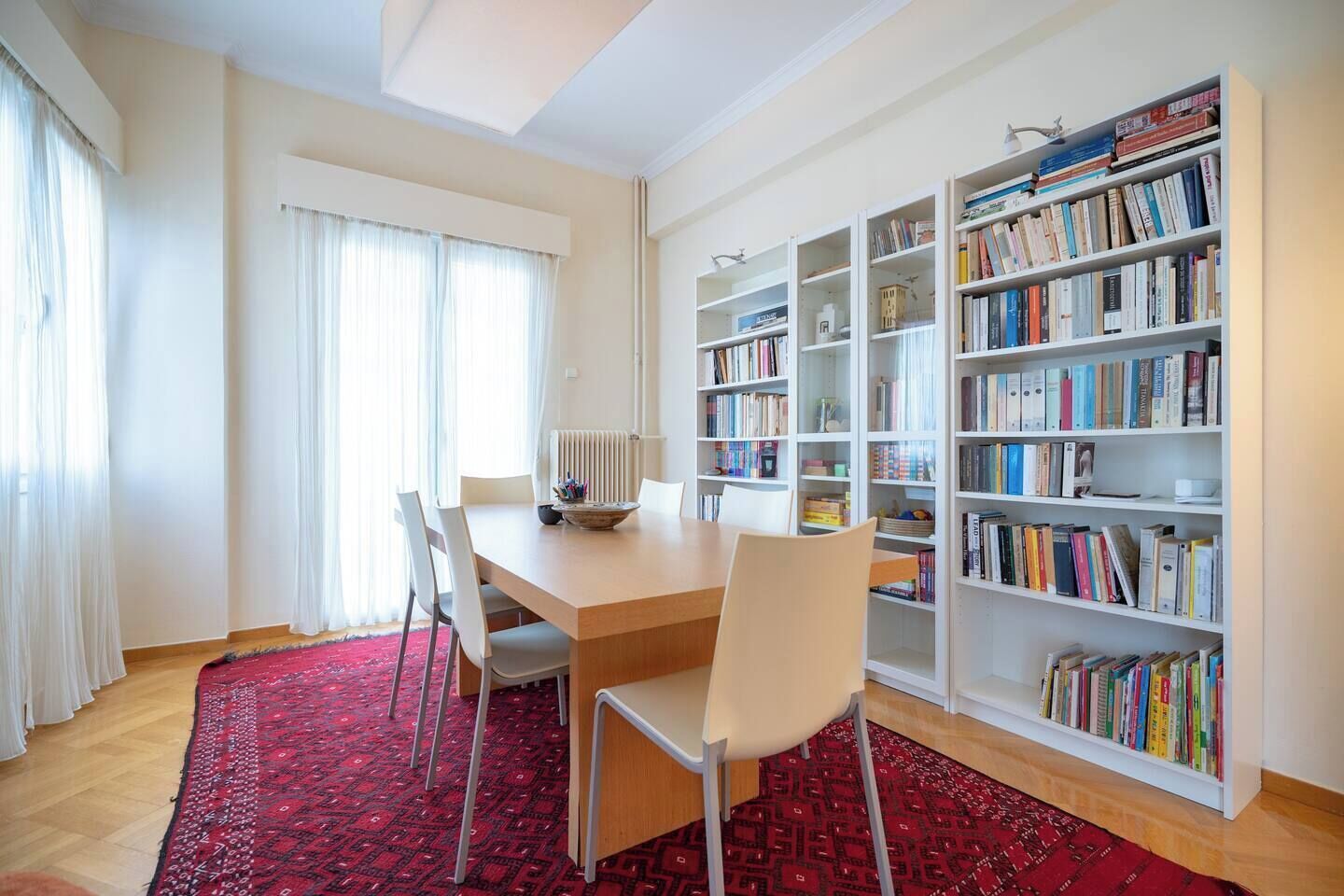 A modern living room with a wooden dining table, white chairs, a red patterned rug, and a large bookshelf filled with books.
