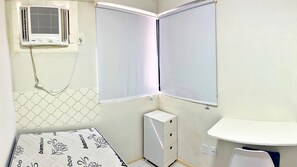 Ein kleiner, sauberer Raum mit einem Bett, einem Nachttisch, einer kleinen Kommode und einem Fenster mit Jalousien.