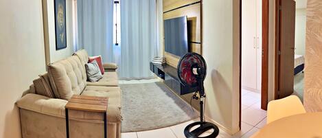 Un salon avec un canapé beige, une petite table d’appoint en bois, un ventilateur sur pied et un téléviseur posé sur un meuble en bois.