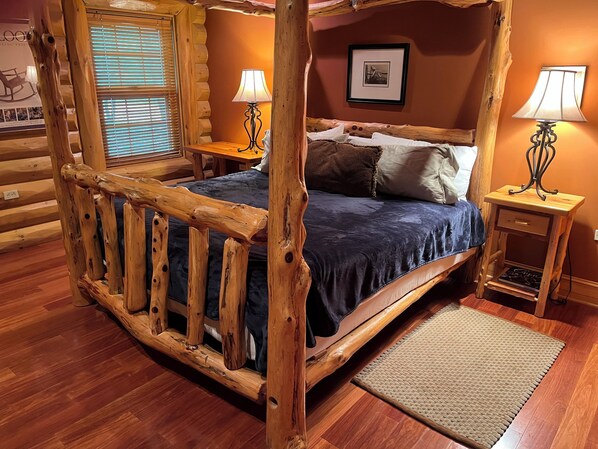 A rustic wooden four-poster bed with a dark blue bedspread and pillows, flanked by two nightstands with lamps, in a room with wooden floors and a window with blinds. - Mystic Pines log home in Shenandoah with hot tub, exercise equipment and views! (Bentonville)