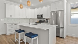 Una cocina moderna con armarios blancos, electrodomésticos de acero inoxidable, una isla grande con taburetes altos e iluminación colgante.