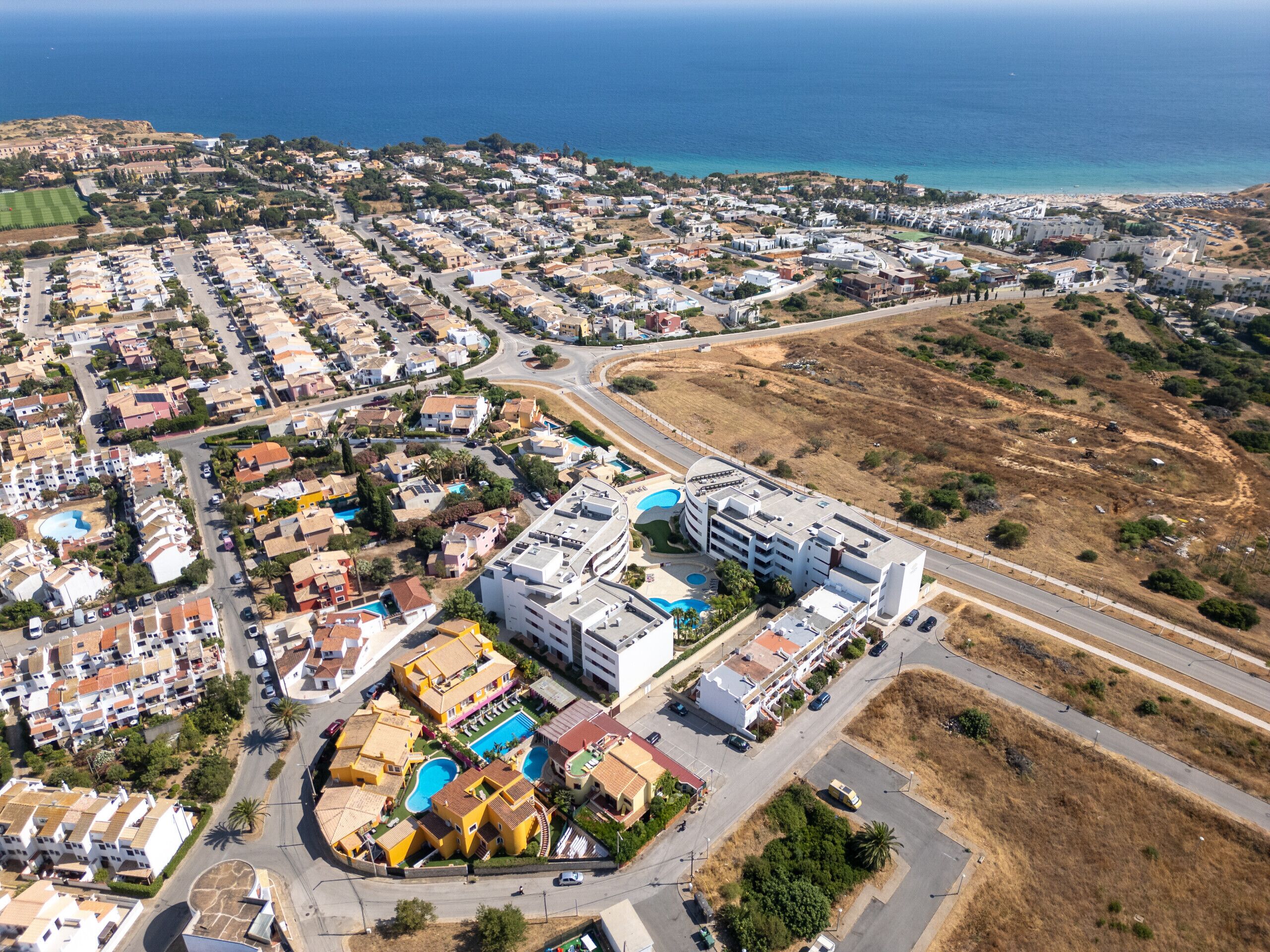 Vista aérea de un pueblo costero con una mezcla de edificios residenciales y comerciales, un hotel destacado con piscina y una carretera que conduce a la playa.
