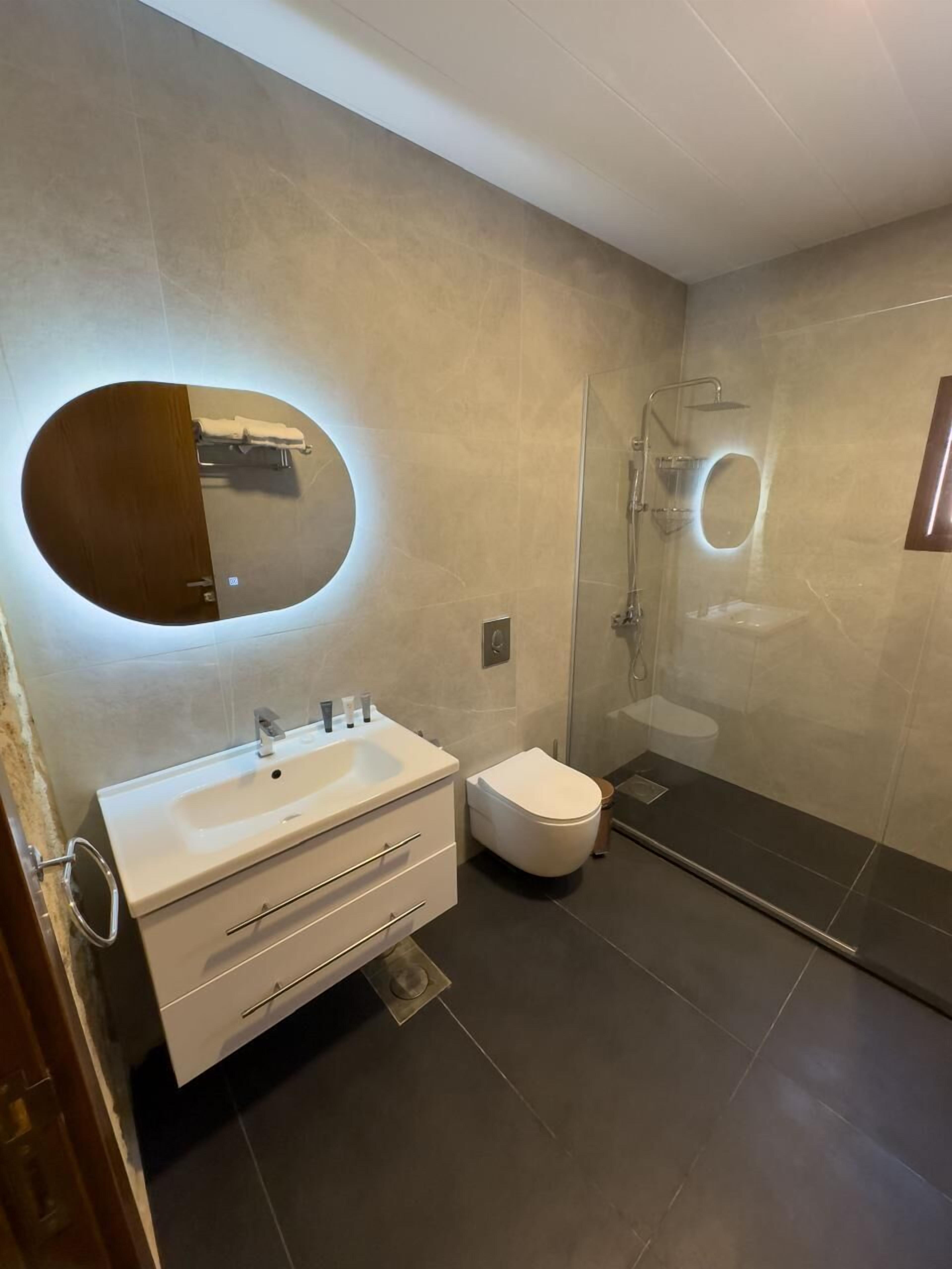 Un baño moderno con un espejo ovalado grande, un lavabo blanco, un inodoro y una ducha con mampara de vidrio.