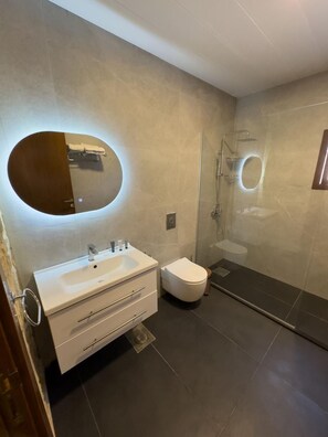 A modern bathroom with a large oval mirror, a white sink, a toilet, and a glass-enclosed shower. - VillaRosa Luxury Boutique (Batroun)