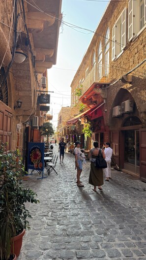 A cobblestone street with buildings on either side, some with balconies and hanging plants. There are signs for a restaurant and a shop. - VillaRosa Luxury Boutique (Batroun)