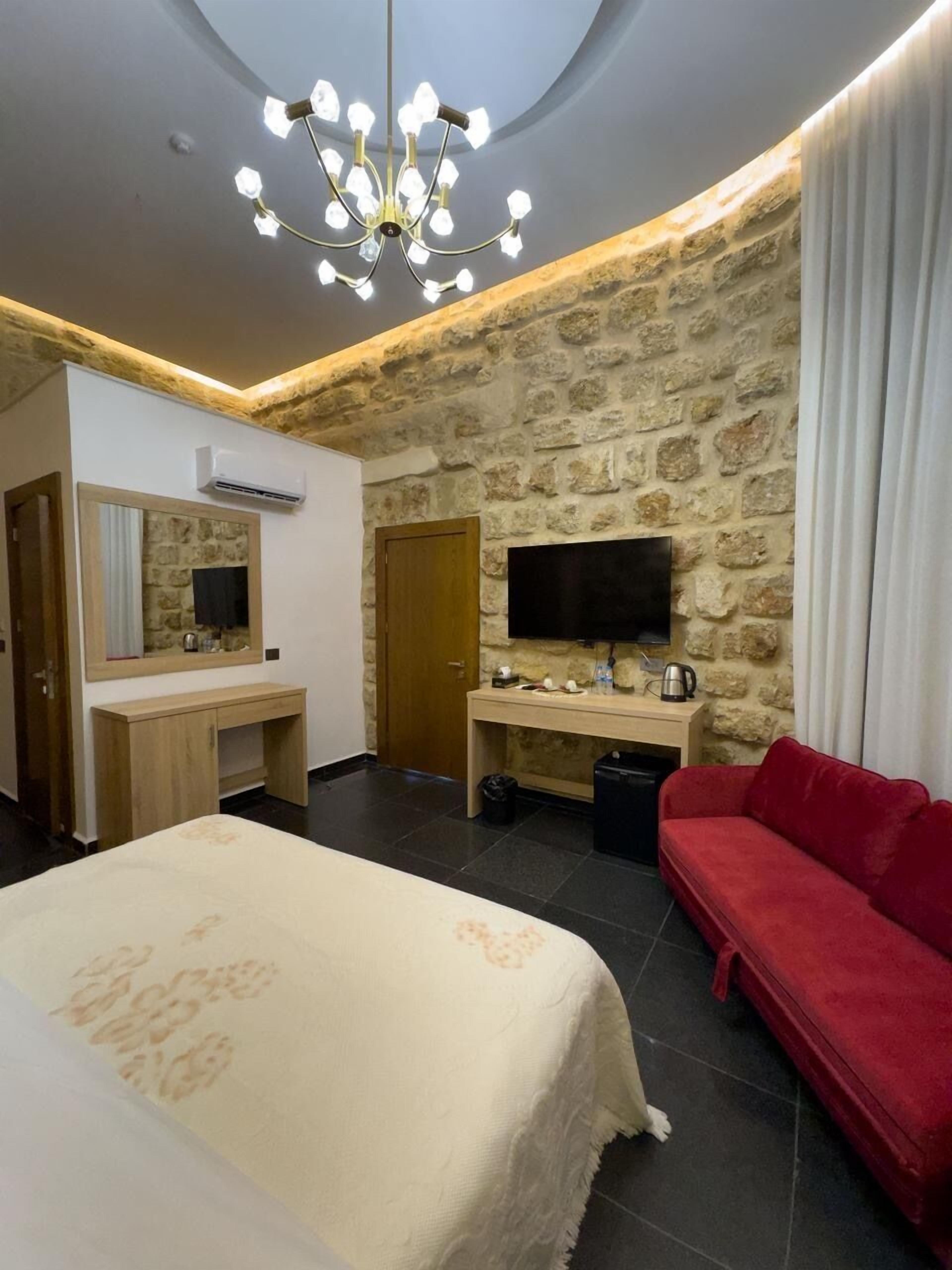 A hotel room with a stone wall, a bed with a white bedspread, a red sofa, a wooden desk, a television, and a chandelier.
