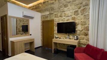 A hotel room with a stone wall, a bed with a white bedspread, a red sofa, a wooden desk, a television, and a chandelier.