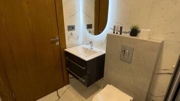 A modern bathroom with a marble floor, a wooden door, a white toilet, a sink with a mirror, and a shower area.